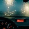 View from car interior with raindrops on windshield and blurred lights at night.